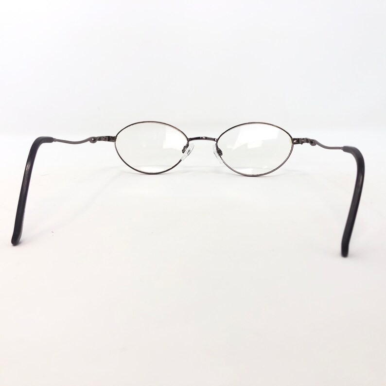 Peut inclure: Lunettes de vue ovales avec une monture en m&eacute;tal fonc&eacute; et des verres transparents. Les branches sont fines et incurv&eacute;es au niveau des branches. Les lunettes sont pos&eacute;es sur un fond blanc.
