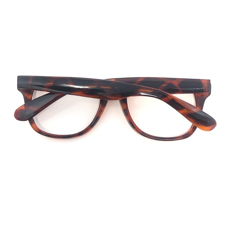 K&ouml;nnte beinhalten: Eine Brille mit Schildpattmuster. Die Fassungen sind eine Mischung aus braunen und bernsteinfarbenen Farbt&ouml;nen mit einer klassischen, leicht abgerundeten Form. Die Brille ist vor einem wei&szlig;en Hintergrund platziert.