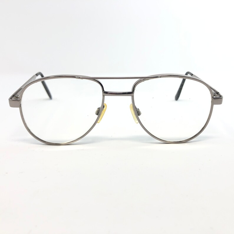 Peut inclure: Une paire de lunettes de style aviateur avec une monture en m&eacute;tal canon de fusil. Les verres sont clairs et les plaquettes nasales sont jaunes. Les lunettes ont un double pont et des branches fines. Le design est &eacute;pur&eacute; et moderne.