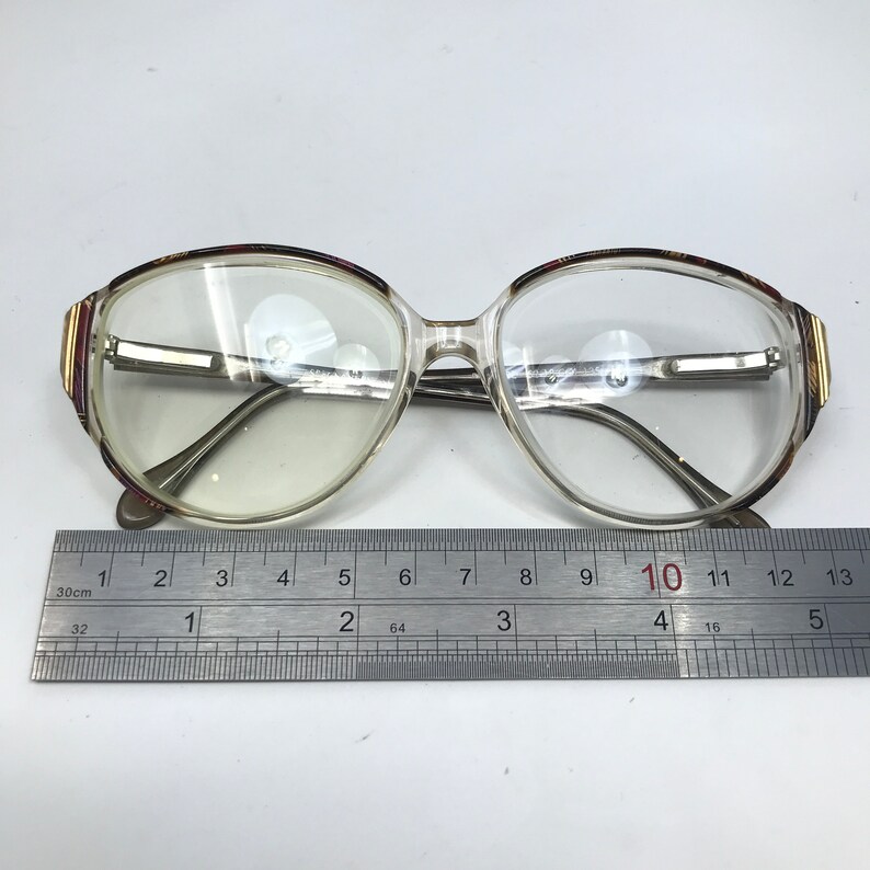 May include: Clear-framed eyeglasses with a rectangular shape, featuring a decorative pattern on the upper corners. The glasses have a silver bridge and arms. A ruler is placed below the glasses, measuring approximately 13 inches.