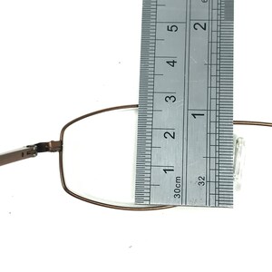 May include: Close-up of a pair of eyeglasses with a brown metal frame. A ruler is placed next to the lens, showing a measurement of approximately 1 inch. The glasses are set against a white background.