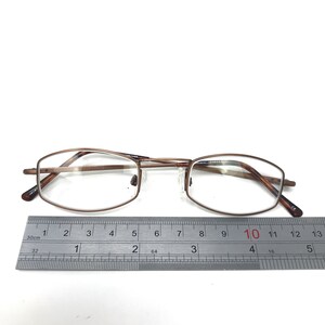 May include: Bronze-colored eyeglasses with rectangular lenses. The glasses are positioned on a white surface next to a ruler, with a measurement of approximately 5 inches.