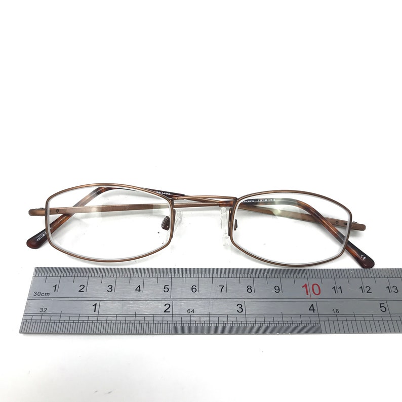 May include: Bronze-colored eyeglasses with rectangular lenses. The glasses are positioned on a white surface next to a ruler, with a measurement of approximately 5 inches.