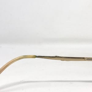 May include: Close-up of a portion of eyeglasses. The frame is a light gold color, with a curved temple arm that transitions from a light beige to a darker green where it connects to the frame. The frame is a thin, rectangular shape.