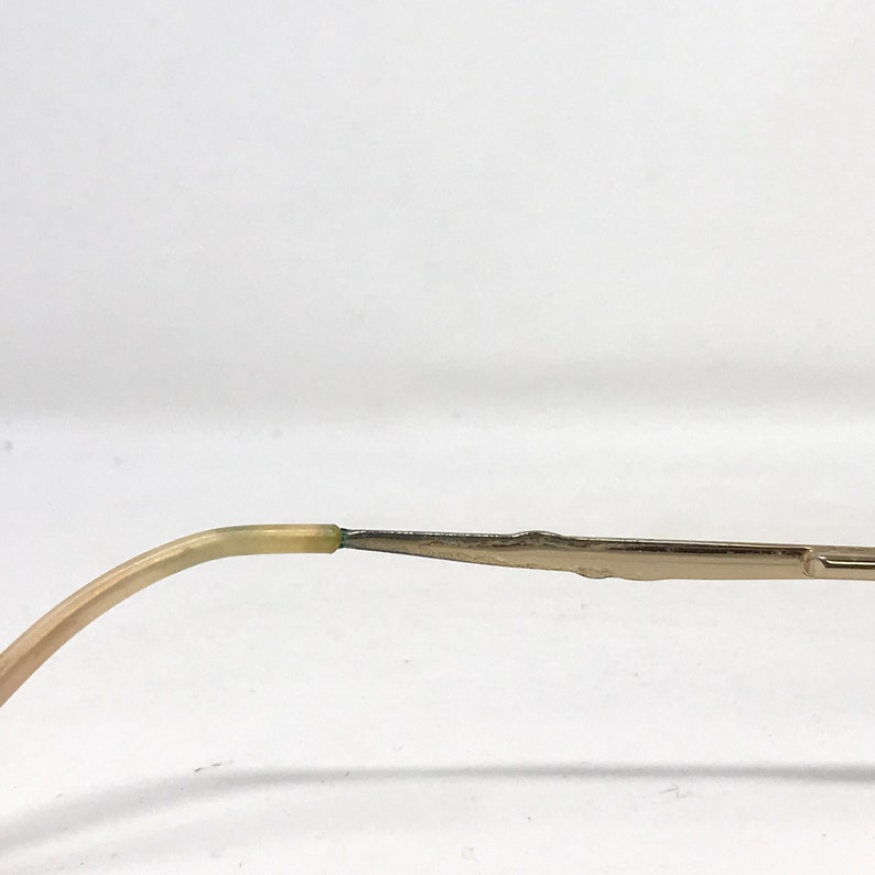 May include: Close-up of a portion of eyeglasses. The frame is a light gold color, with a curved temple arm that transitions from a light beige to a darker green where it connects to the frame. The frame is a thin, rectangular shape.