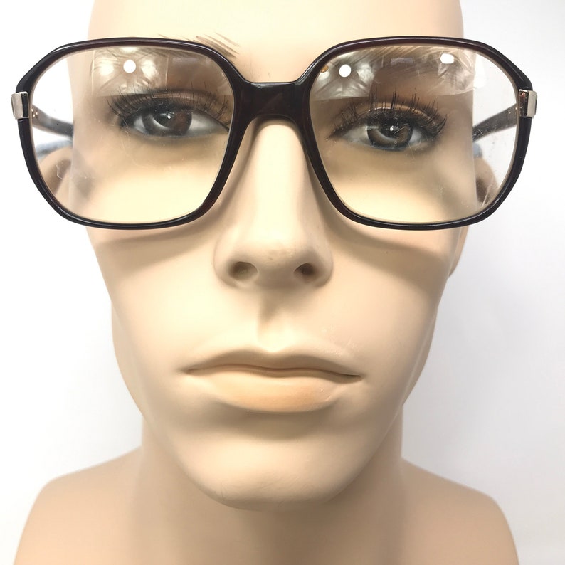 May include: Dark brown, square-framed eyeglasses. The glasses have clear lenses and silver-coloured accents on the temples. The glasses are displayed on a mannequin.