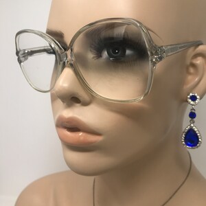May include: Clear, oversized eyeglasses with a square shape are featured. The glasses are on a mannequin. Dangling earrings with blue teardrop-shaped gems and silver accents are also visible. The mannequin has a neutral skin tone and is set against a white background.