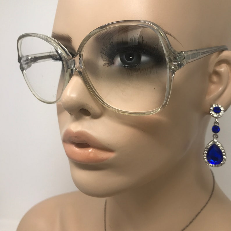 May include: Clear, oversized eyeglasses with a square shape are featured. The glasses are on a mannequin. Dangling earrings with blue teardrop-shaped gems and silver accents are also visible. The mannequin has a neutral skin tone and is set against a white background.