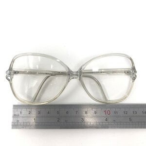 May include: Clear, oversized eyeglasses with a butterfly-shaped frame. The glasses have the text "SPECSAVERS 1753156" and "DELLA 21024207" printed on the frame. A ruler is placed below the glasses, measuring approximately 5 inches in length.