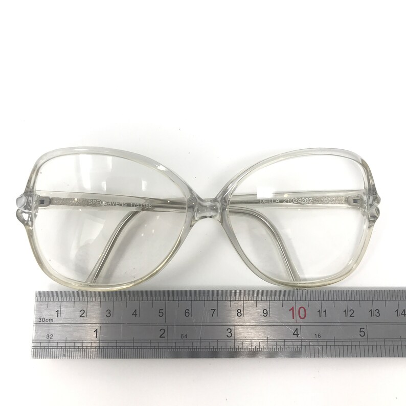 May include: Clear, oversized eyeglasses with a butterfly-shaped frame. The glasses have the text "SPECSAVERS 1753156" and "DELLA 21024207" printed on the frame. A ruler is placed below the glasses, measuring approximately 5 inches in length.