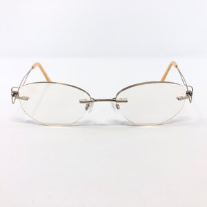 May include: Rimless eyeglasses with oval lenses and a silver-tone metal frame. The temples are a light tan color. The glasses are set against a white background.