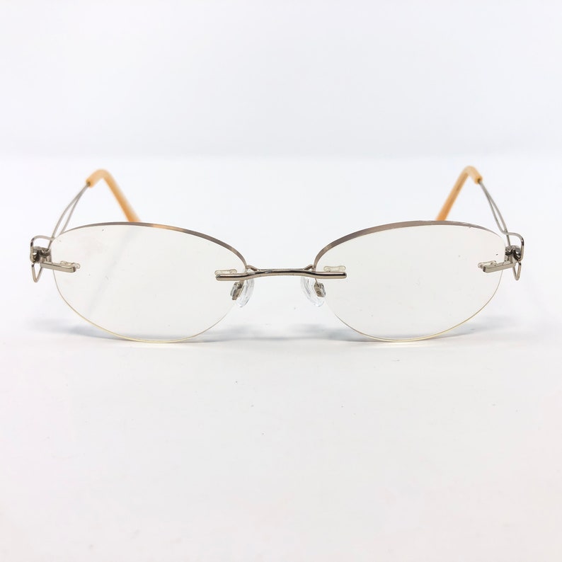 May include: Rimless eyeglasses with oval lenses and a silver-tone metal frame. The temples are a light tan color. The glasses are set against a white background.
