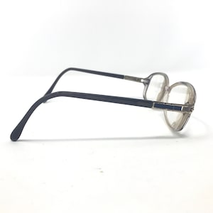 May include: A pair of eyeglasses with a clear, rounded frame and dark gray temples. The lenses are clear, and the frame has a subtle, rectangular detail on the front. The glasses are set against a white background.