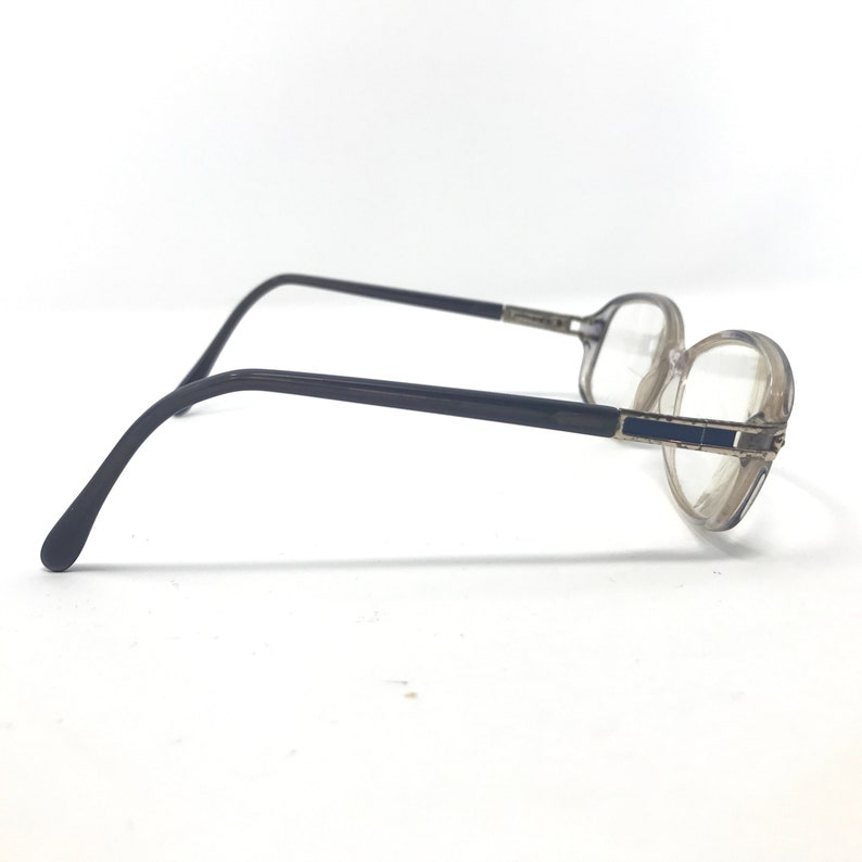 May include: A pair of eyeglasses with a clear, rounded frame and dark gray temples. The lenses are clear, and the frame has a subtle, rectangular detail on the front. The glasses are set against a white background.