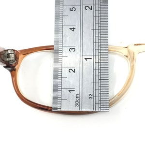 May include: A close-up of a pair of brown and beige eyeglasses with a ruler measuring the width of the frame at 1 3/8 inches.