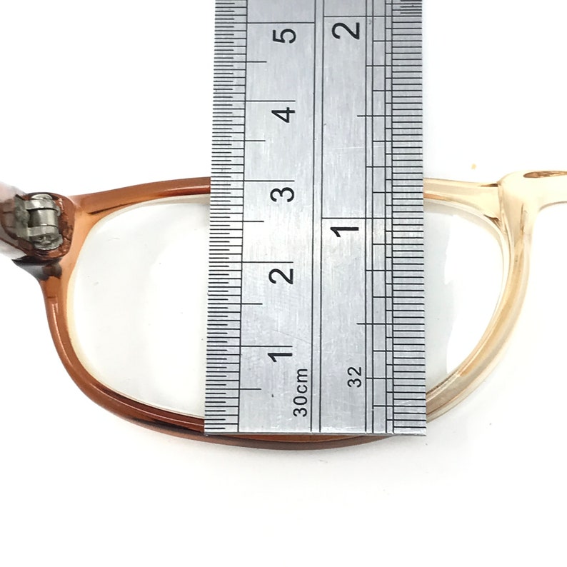 May include: A close-up of a pair of brown and beige eyeglasses with a ruler measuring the width of the frame at 1 3/8 inches.