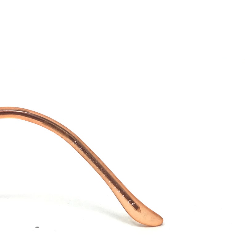 May include: Close-up of a copper-colored eyeglass frame arm. The arm curves gently and ends in a rounded tip. The frame appears to be made of metal and has a smooth, reflective surface. The background is white.