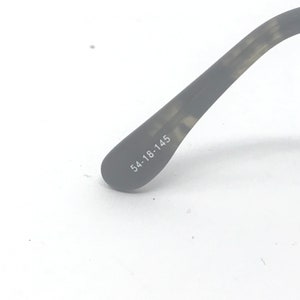 Puede incluir: Primer plano de una parte de gafas. La patilla gris oscuro de las gafas es visible, con las medidas "54-18-145" impresas en blanco. La patilla de las gafas tiene un patr&oacute;n texturizado sobre un fondo blanco.