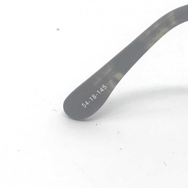 Puede incluir: Primer plano de una parte de gafas. La patilla gris oscuro de las gafas es visible, con las medidas "54-18-145" impresas en blanco. La patilla de las gafas tiene un patr&oacute;n texturizado sobre un fondo blanco.
