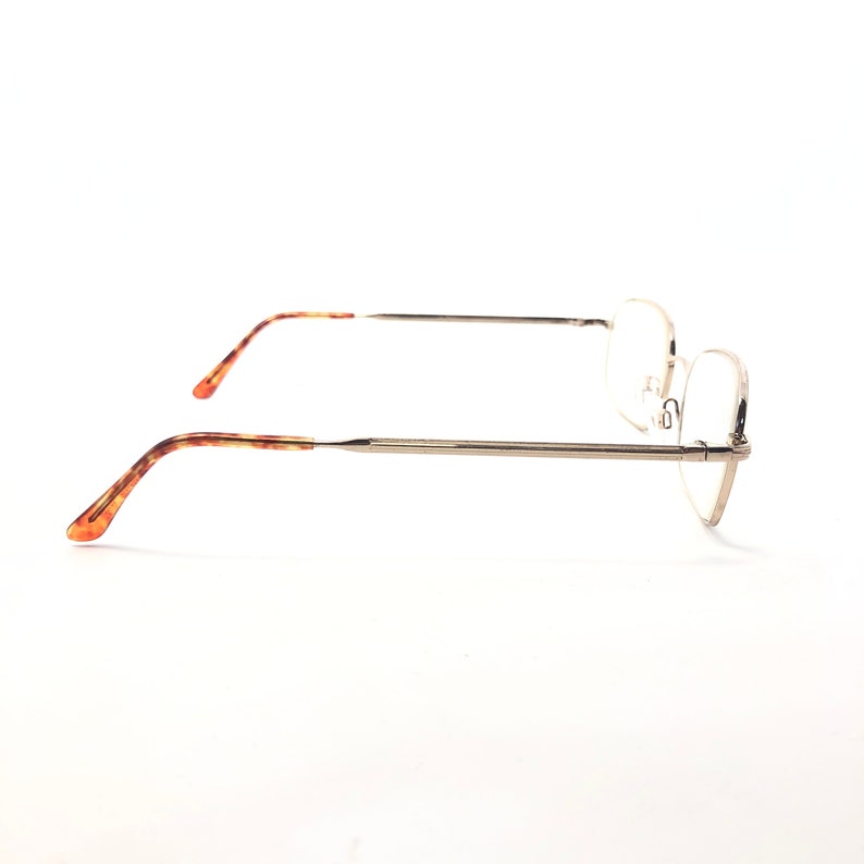 May include: Eyeglasses with a silver metal frame and clear lenses. The temples feature a tortoiseshell pattern in brown and orange. The glasses are set against a white background.