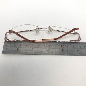 May include: Rimless eyeglasses with a brown metal frame. The glasses are measured with a ruler, showing a length of approximately 5 inches. The lenses are clear, and the nose pads are transparent.