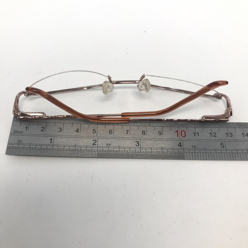 May include: Rimless eyeglasses with a brown metal frame. The glasses are measured with a ruler, showing a length of approximately 5 inches. The lenses are clear, and the nose pads are transparent.