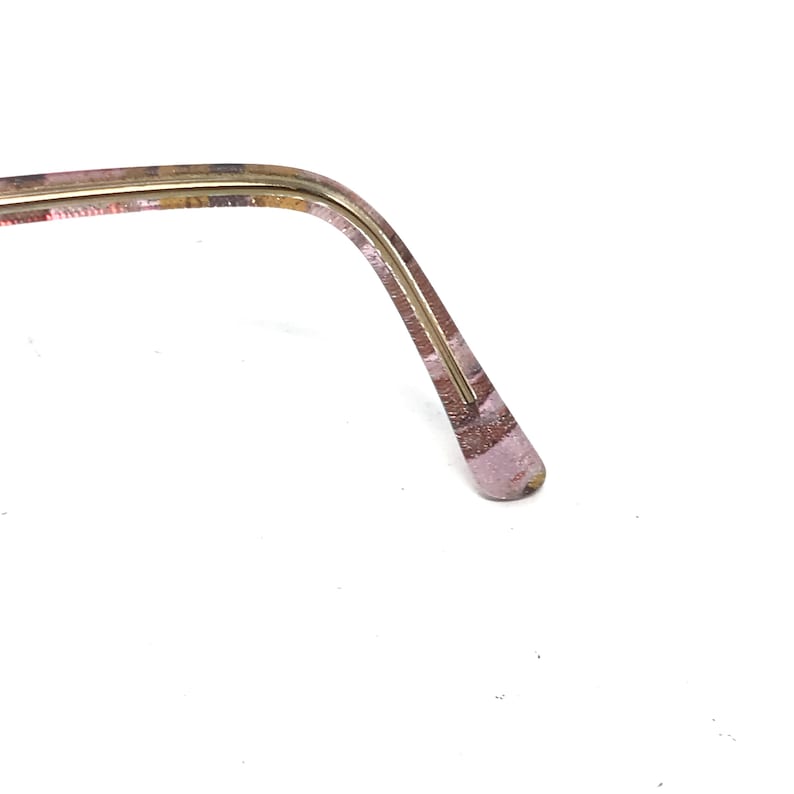 May include: Close-up of the temple arm of eyeglasses. The arm is a light pink color with a speckled pattern and a gold-colored metal frame. The glasses are set against a white background.