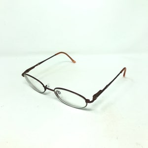 May include: Oval-shaped eyeglasses with a dark brown metal frame and clear lenses. The temples and earpieces are a lighter brown. The glasses are set against a white background.