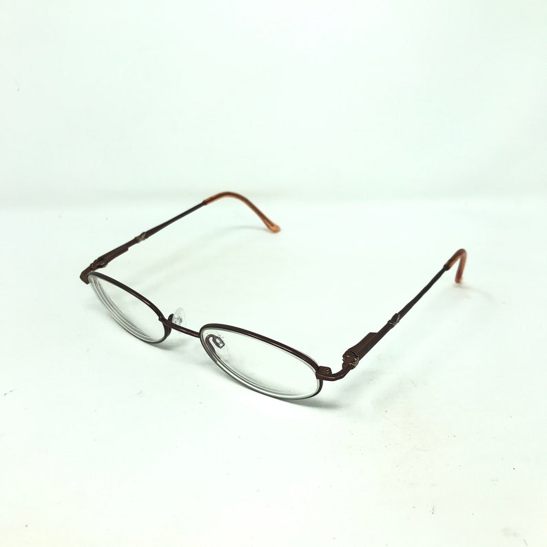 May include: Oval-shaped eyeglasses with a dark brown metal frame and clear lenses. The temples and earpieces are a lighter brown. The glasses are set against a white background.