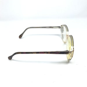 May include: A pair of eyeglasses with round lenses and thin, silver-toned frames. The temples feature a decorative pattern in shades of brown, burgundy, and gold. The glasses are set against a white background, showcasing their design.
