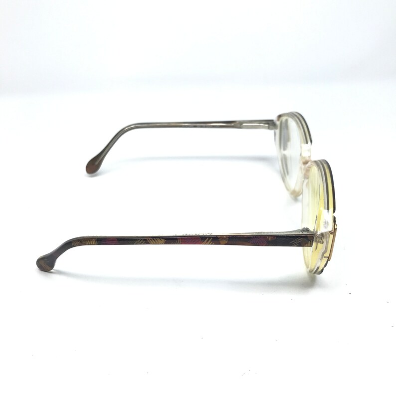 May include: A pair of eyeglasses with round lenses and thin, silver-toned frames. The temples feature a decorative pattern in shades of brown, burgundy, and gold. The glasses are set against a white background, showcasing their design.