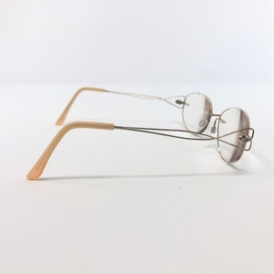 May include: A pair of eyeglasses with a thin, silver-colored metal frame and round lenses. The earpieces are a light beige color. The glasses are set against a plain white background.