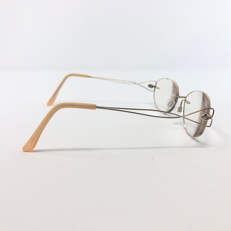 May include: A pair of eyeglasses with a thin, silver-colored metal frame and round lenses. The earpieces are a light beige color. The glasses are set against a plain white background.