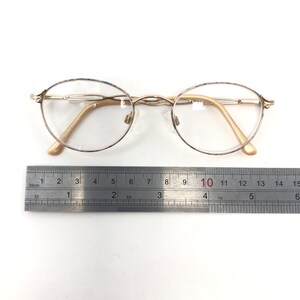 May include: Gold-toned oval eyeglasses with clear lenses. The glasses have a delicate frame and nose pads. A ruler is placed next to the glasses, showing measurements in inches. The glasses are on a white background.