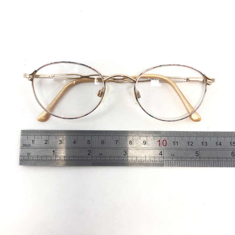 May include: Gold-toned oval eyeglasses with clear lenses. The glasses have a delicate frame and nose pads. A ruler is placed next to the glasses, showing measurements in inches. The glasses are on a white background.