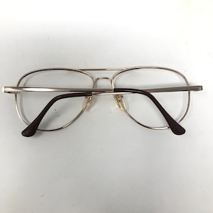 May include: Silver-toned aviator eyeglasses with clear lenses and brown temples. The glasses have a double bridge and a classic, retro design. The lenses are round and the frame is thin and metallic.
