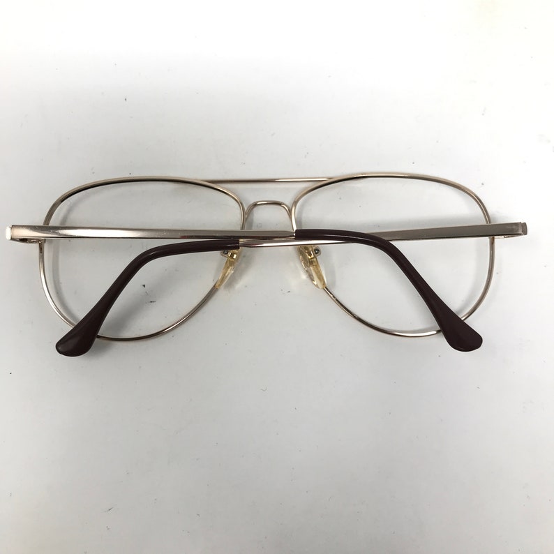 May include: Silver-toned aviator eyeglasses with clear lenses and brown temples. The glasses have a double bridge and a classic, retro design. The lenses are round and the frame is thin and metallic.