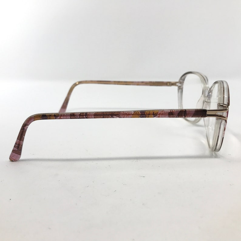 May include: Clear eyeglasses with a round frame and patterned temples. The temples feature a pink, brown, and gold floral design. The glasses are viewed from the side against a white background.