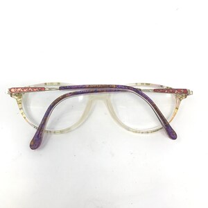 May include: Clear eyeglasses with a light-colored frame and purple temples. The frame has a decorative design in pink and gold. The lenses are clear, and the glasses are set against a white background.