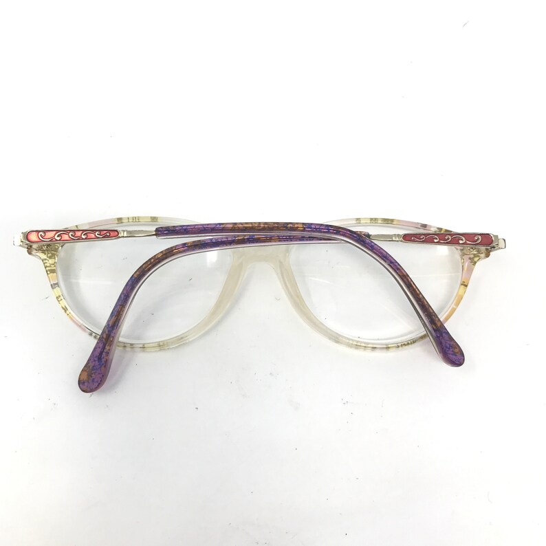 May include: Clear eyeglasses with a light-colored frame and purple temples. The frame has a decorative design in pink and gold. The lenses are clear, and the glasses are set against a white background.