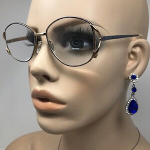 Op de afbeelding: Een bril met gouden montuur en een blauw accent op de veren en heldere glazen. De bril wordt gecombineerd met bungelende oorbellen met een blauwe druppelvormige edelsteen omringd door kleinere heldere stenen. De oorbellen hebben een zilverkleurige zetting.