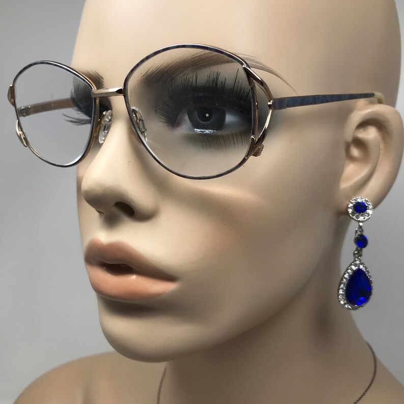 Op de afbeelding: Een bril met gouden montuur en een blauw accent op de veren en heldere glazen. De bril wordt gecombineerd met bungelende oorbellen met een blauwe druppelvormige edelsteen omringd door kleinere heldere stenen. De oorbellen hebben een zilverkleurige zetting.
