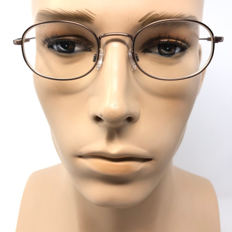 Puede incluir: Primer plano de gafas con montura ovalada de metal marr&oacute;n. Las gafas se muestran sobre una cabeza de maniqu&iacute;. La montura tiene un dise&ntilde;o fino y delicado. Fondo blanco.