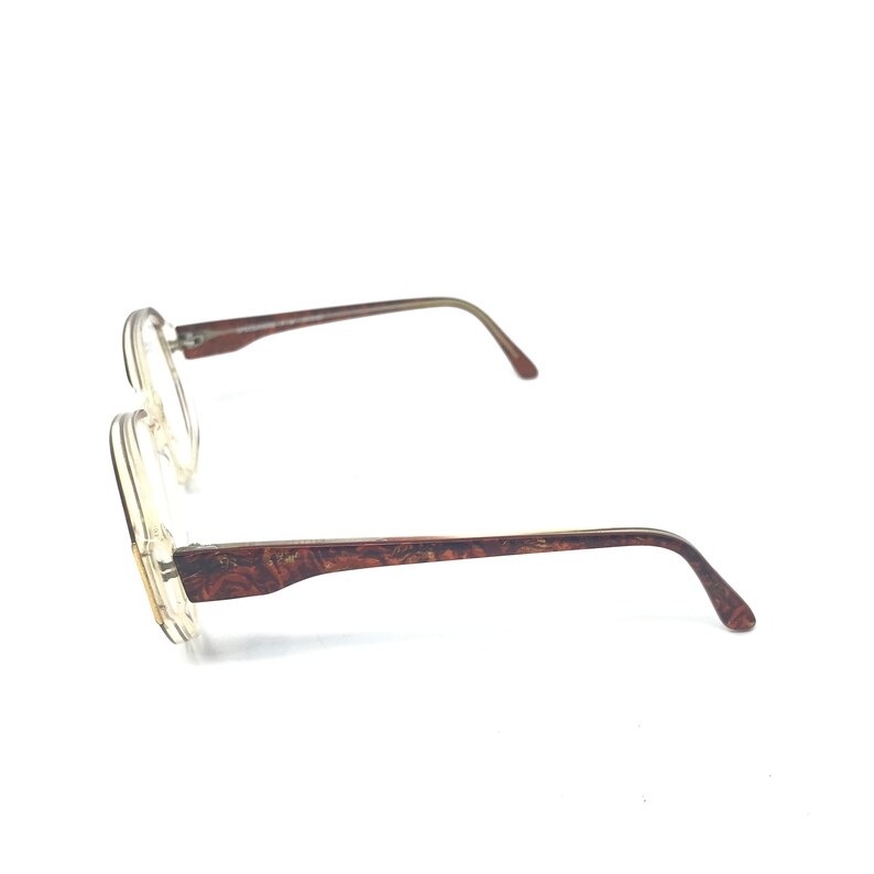 May include: Clear-framed eyeglasses with brown patterned temples and thin, brown arms. The lenses are round, and the frames are a light, transparent color. The glasses are angled, showing the side view.