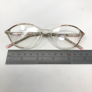 May include: Clear eyeglasses with oval lenses and patterned temples. The frames are transparent, and the temples have a floral design in shades of pink, red, and brown. A ruler is placed below the glasses, measuring approximately 5 inches.