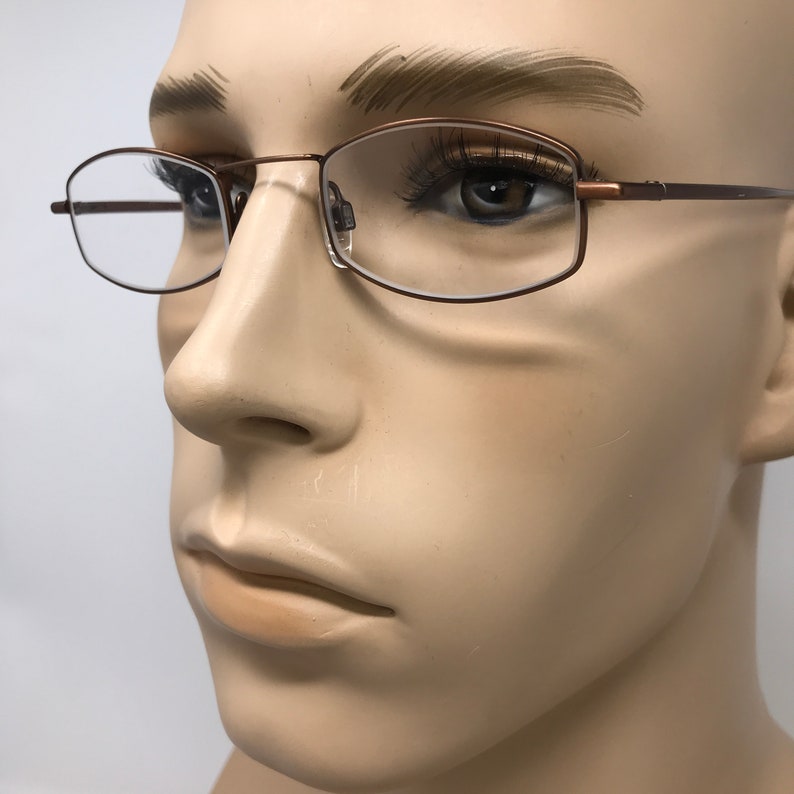 May include: Bronze-colored eyeglasses with a rectangular frame and clear lenses. The glasses are set against a neutral background, showcasing the design and details of the eyewear. The frames have a thin, delicate appearance.
