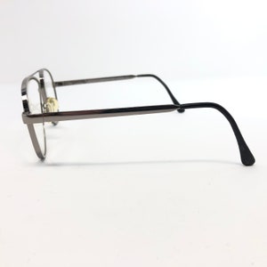 Peut inclure: Une paire de lunettes avec une monture en m&eacute;tal gris fonc&eacute; et des verres transparents. Les lunettes ont une forme arrondie et des branches droites qui se courbent vers le bas aux extr&eacute;mit&eacute;s. Sur fond blanc.