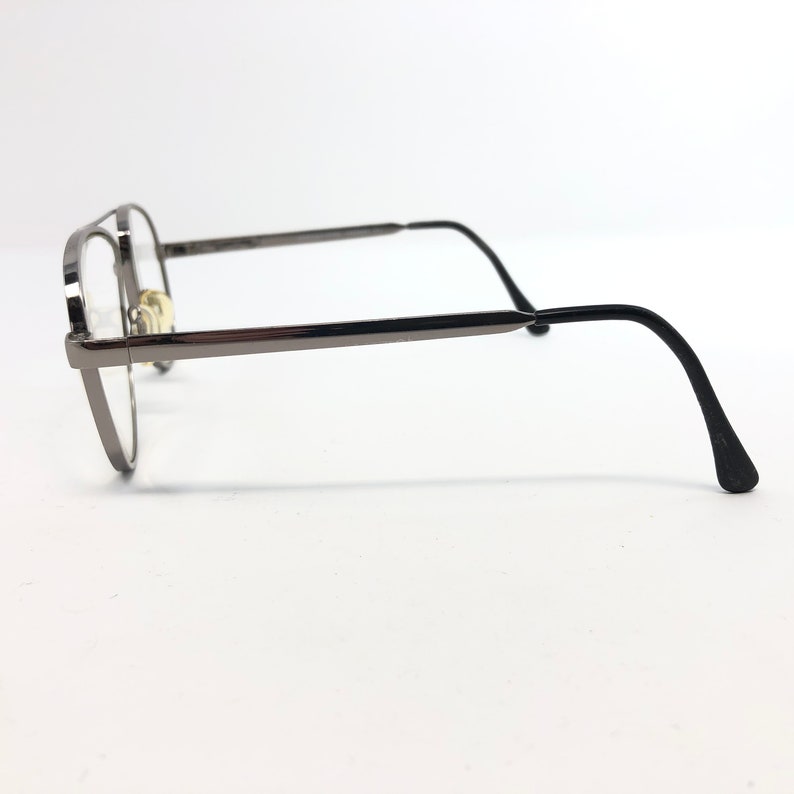 Peut inclure: Une paire de lunettes avec une monture en m&eacute;tal gris fonc&eacute; et des verres transparents. Les lunettes ont une forme arrondie et des branches droites qui se courbent vers le bas aux extr&eacute;mit&eacute;s. Sur fond blanc.
