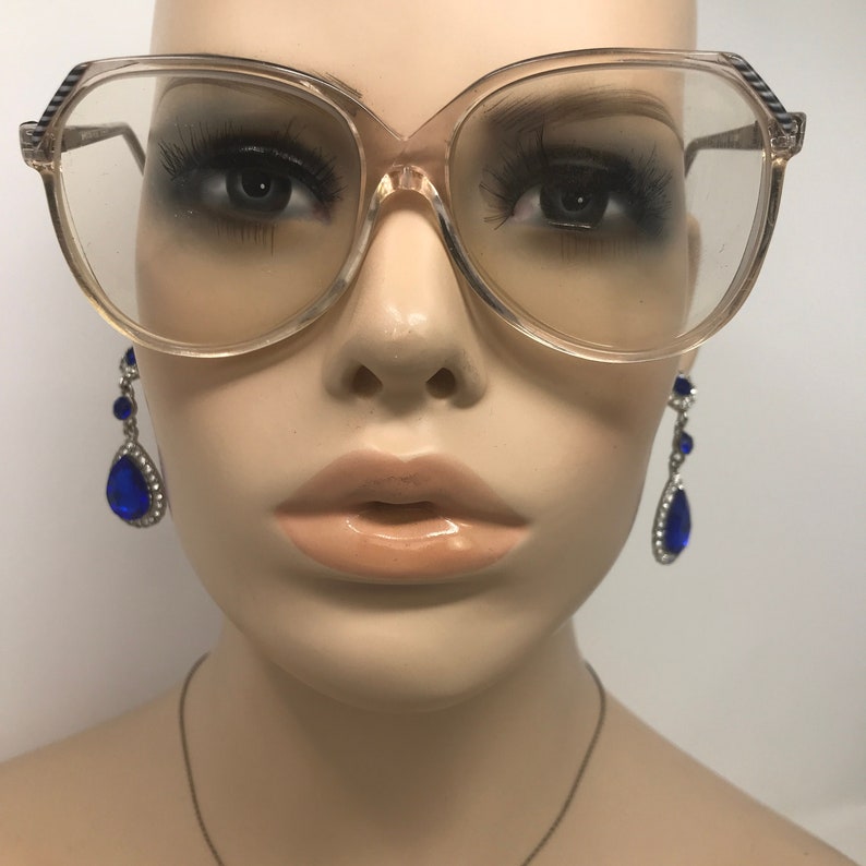 May include: Clear, oversized eyeglasses with a light brown frame and black and white striped accents on the temples. The mannequin is also wearing blue teardrop earrings with silver accents.