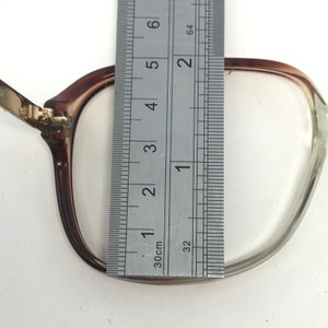 Op de afbeelding: Close-up van een bril met bruine monturen en heldere glazen. Een metalen liniaal is over de bril geplaatst, met een breedte van ongeveer 5 cm. De bril ligt op een witte ondergrond.